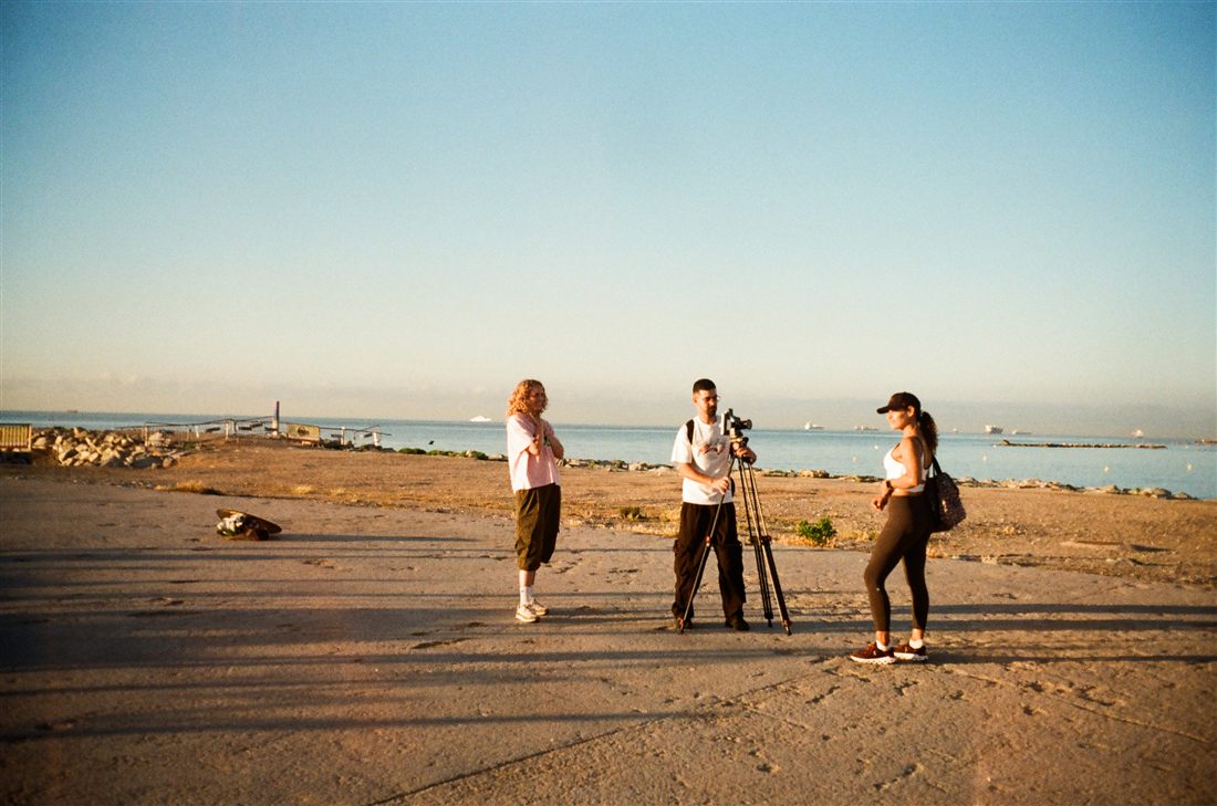 Three people filming on a concrete waterfront at golden hour, one behind a camera on a tripod, a skateboard on the ground and a breakwater in the sea behind them.